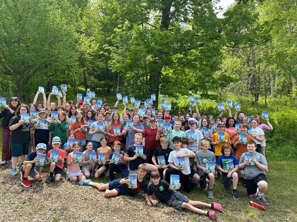 Large group of about 80 6th graders standing together for  a photo in a wooded field, holding copies of THE TROUBLE WITH HEROES 