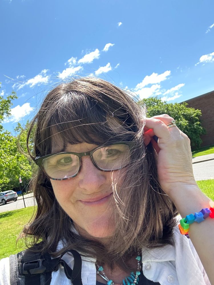 Author Kate Messner outside a school building on a sunny blue-sky day, wearing a white shirt & homemade rainbow bracelet.