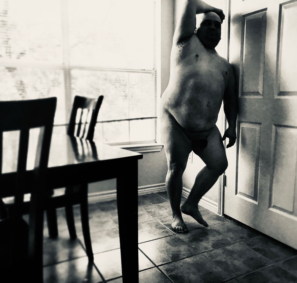 Black-and-white photograph of an adult man standing nude in a sunlit room, leaning against a door with one arm raised above his head. Strong window light casts dramatic shadows across his body, with a table and chairs visible in the foreground on a tiled floor.