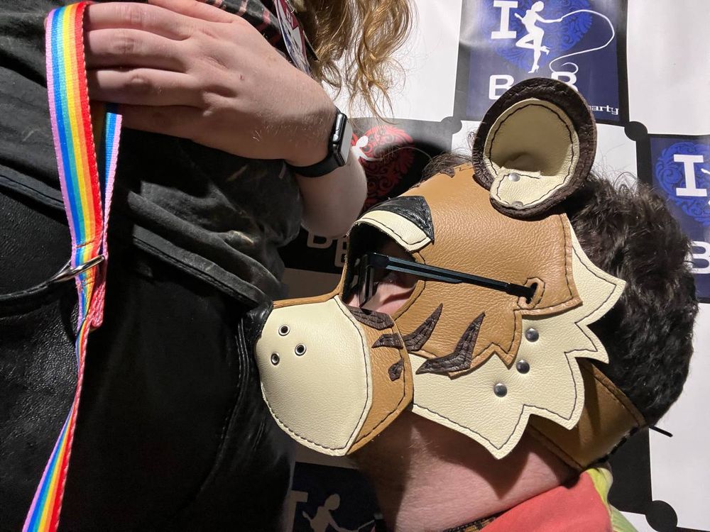 a side view of me in a tiger pup hood with my muzzle against my partner’s leather shorts. he’s holding a rainbow patterned leash