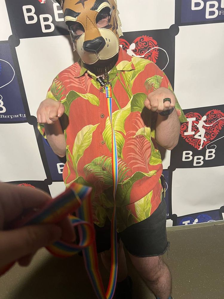 me in a pup hood and pink shirt with green leaves, holding my hands out like paws. the photographer (my partner) is holding a rainbow leash attached to my collar