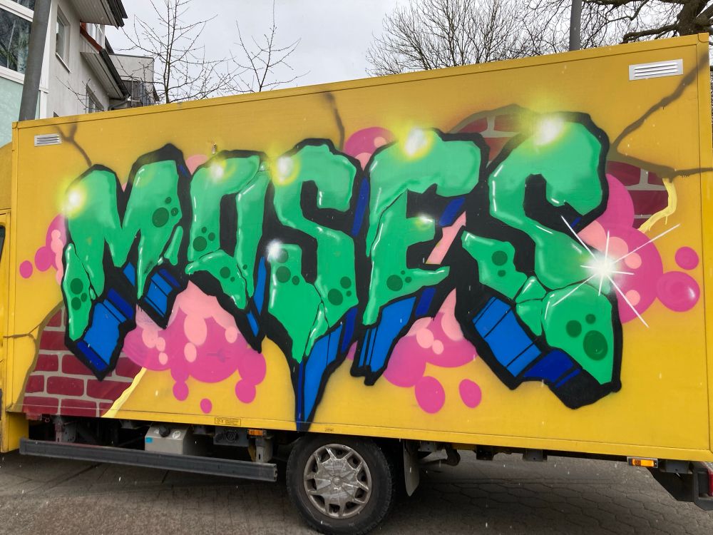 The image features a colorful graffiti mural on a yellow truck, prominently displaying the name "MOSES" in large, stylized letters, looking like stone. The letters are green with splashes of blue and pink accents, set against a background that includes a brick pattern and is surrounded by pink bubble-like shapes. The wide shadow is blue and looks also like stone.