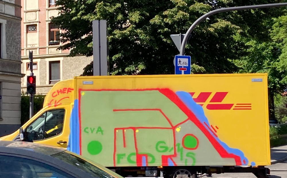 A graffiti-covered yellow delivery truck is parked at a traffic light. The side of the truck features colorful abstract designs and text, including the word "CHEF“, which is written above the drivers cabin and an arrow points to the seat. The huge graffiti is sage green with red and blue outlines and Drop shadow. The word „FUTO“ starts with the letter F and the strokes of the F are like a roof for the other letters placed below. Some brighter green dots and letters are spread over the lower part of the big letters.
 In the background, traffic signals and road signs can be seen, along with trees and nearby
