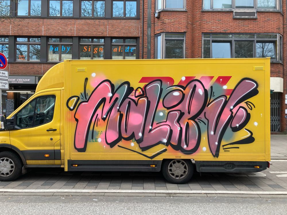 DHL-truck with multicoloured graffiti on the side. Looks like the word „MaLiBU“ to me. Black outlined letters with some lines indicating movement and a slight drop shadow to the bottom right. The M is mostly pink, the following letters are in an old-pink/violet, dotted with bright pink and some greenish Color in the background. Sind Highlights add white dots are sprinkled over the whole piece.