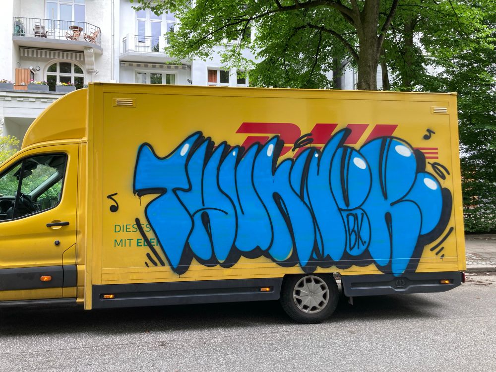 A yellow delivery truck with a large blue graffiti tag on its side that reads "THUNDER“ is parking on a street. The graffiti features stylized, rounded letters with additional markings. The background includes a building with balconies and green trees.