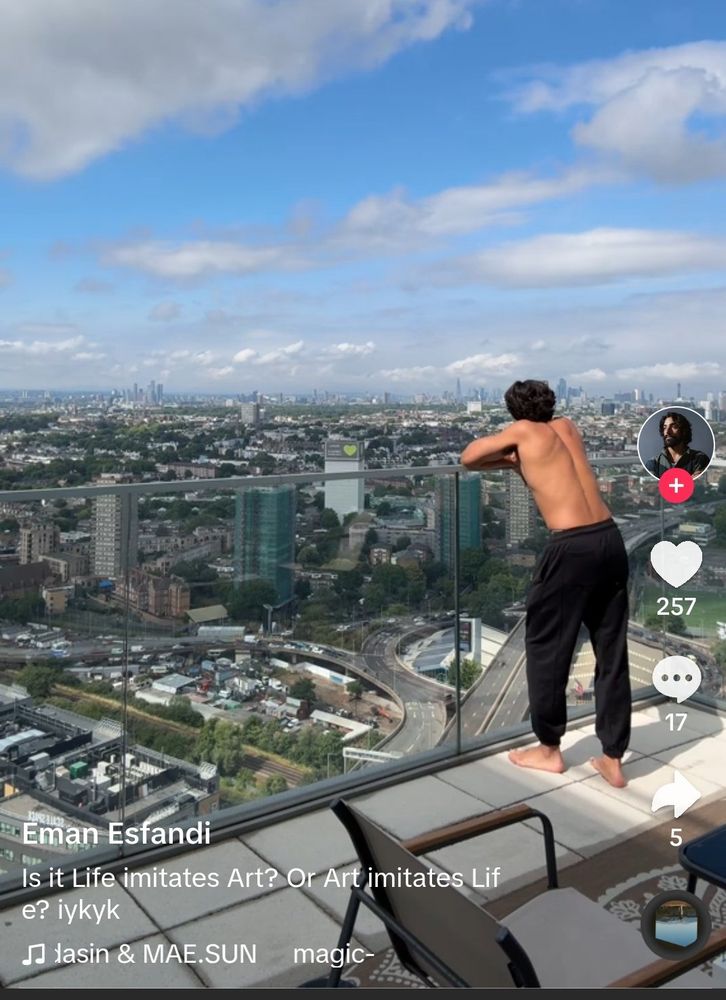 Eman Esfandi stands on top of a tall building and leans against a glass railing looking out at a vast cityscape. He is standing just like Ezra Bridger does in Star Wars Rebels. 