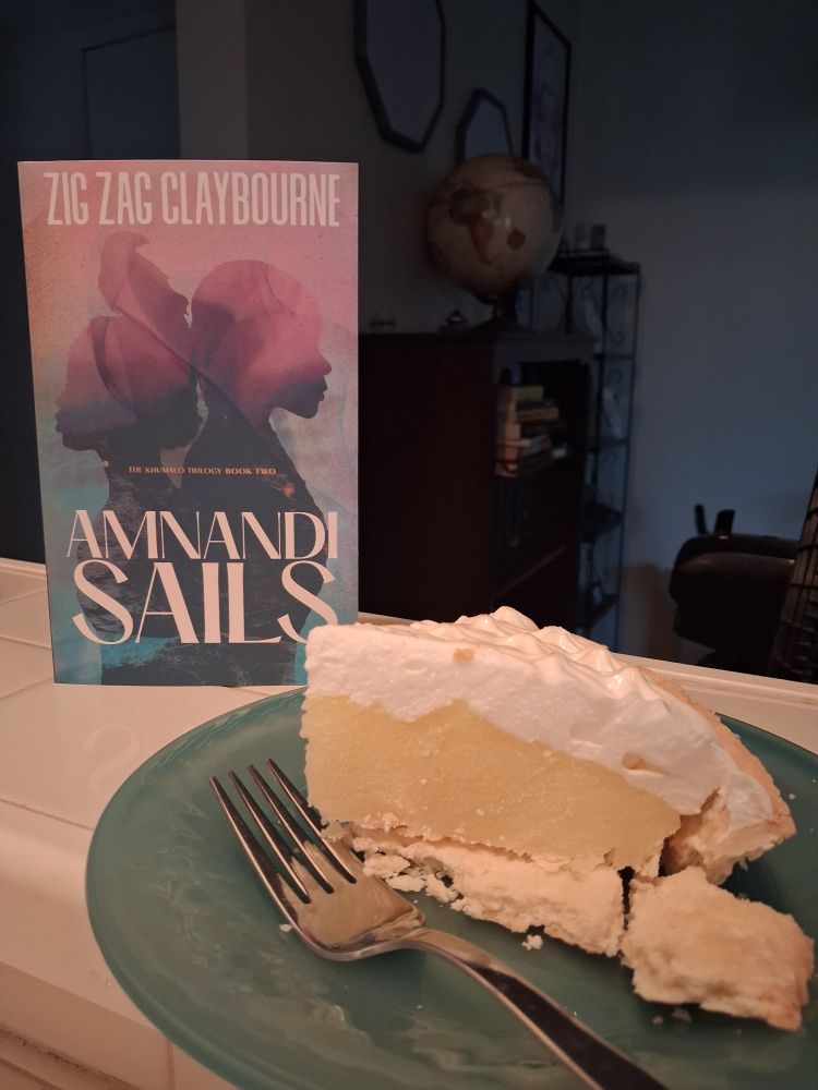 The book AMNANDI SAILS stands at attention behind a large slice of lemon meringue pie sitting on a foam-blue plastic saucer with a silver fork perfectly nearby.