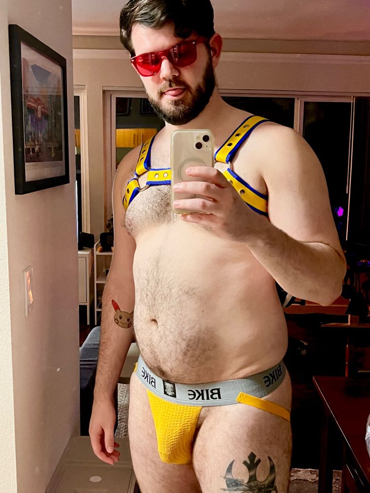 Man dressed in a yellow and blue chest harness, a yellow jock strap and red glasses. 