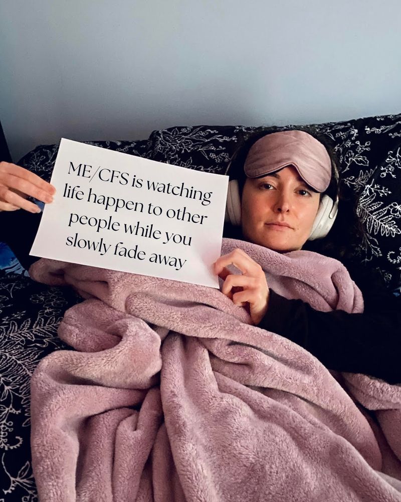 Samatha is holding a sign that reads: "ME/CFS is watching life happen to other people while you slowly fade away.