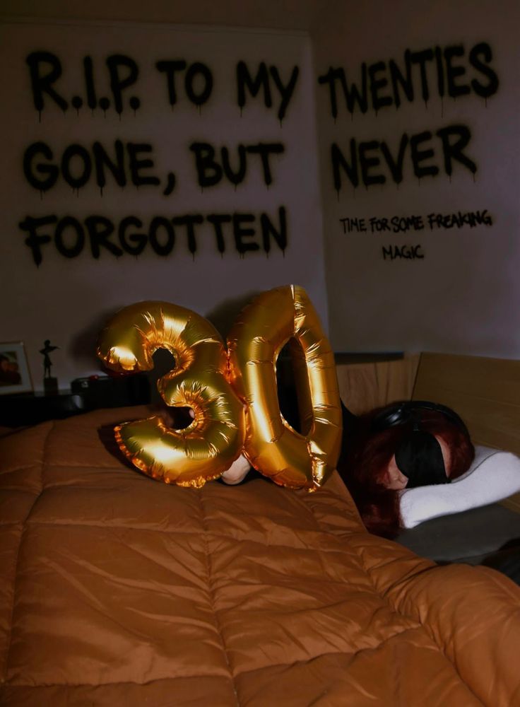 In a dark room, golden foil balloons shaped as the number "30" rest on top of a brown comforter. Behind the balloons, Bente—a person living with very severe ME—is lying in bed, wearing a sleep mask and noise-canceling headphones.The wall behind her features bold, black text that reads: 'R.I.P. TO MY TWENTIES — GONE, BUT NEVER FORGOTTEN’.