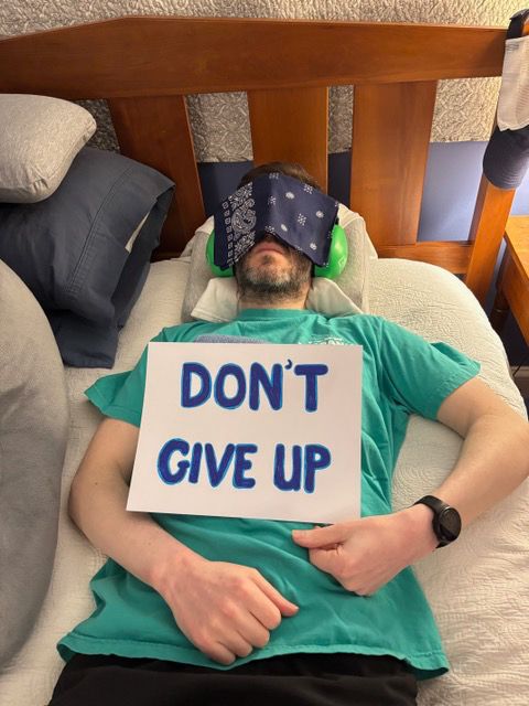 Brent is lying in bed with a cover over his eyes. A sign on his chest reads, 'Don't give up.'