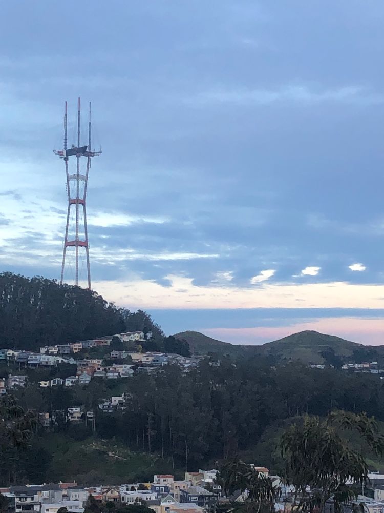 Image description: Sutro tower as seen from the west at dusk, with clouds behind and houses below 