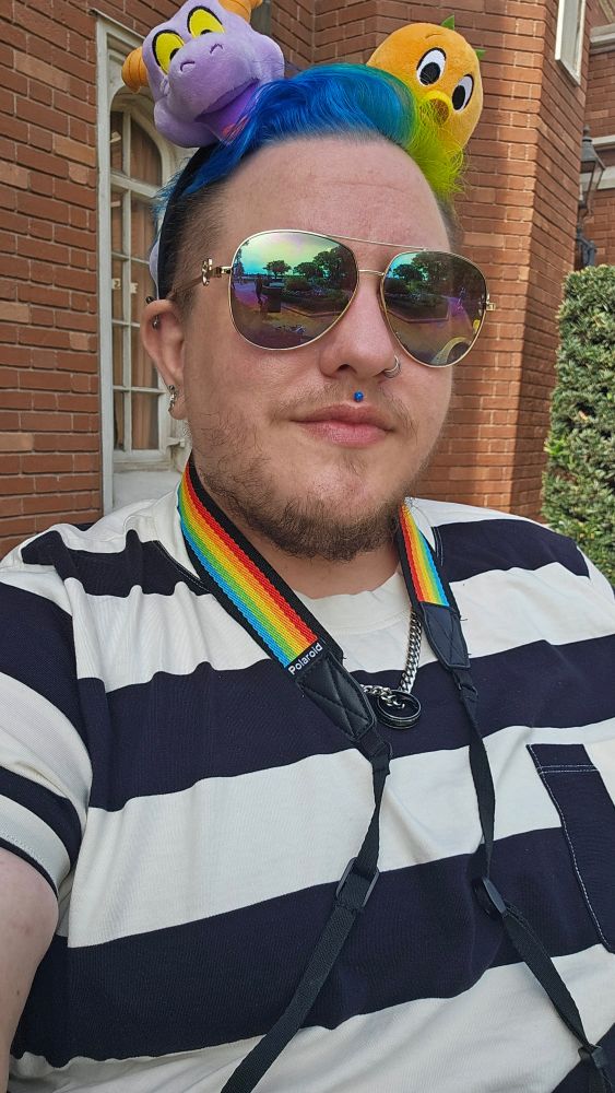 KC, a fat white human with a brown beard and blue and highlighter green hair. They're wearing a tshirt with thick black and white stripes, sunglasses, and a headband with orange bird and Figment 