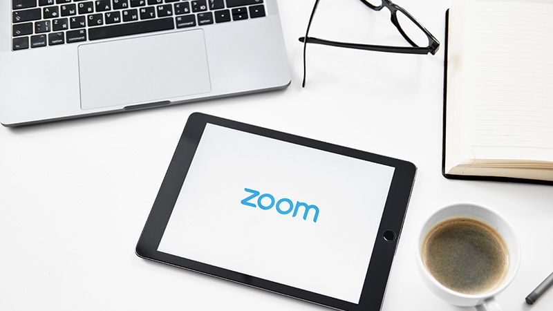 Image of open laptop, tablet displaying the Zoom logo, coffee cup, open book, spectacles on a desk
