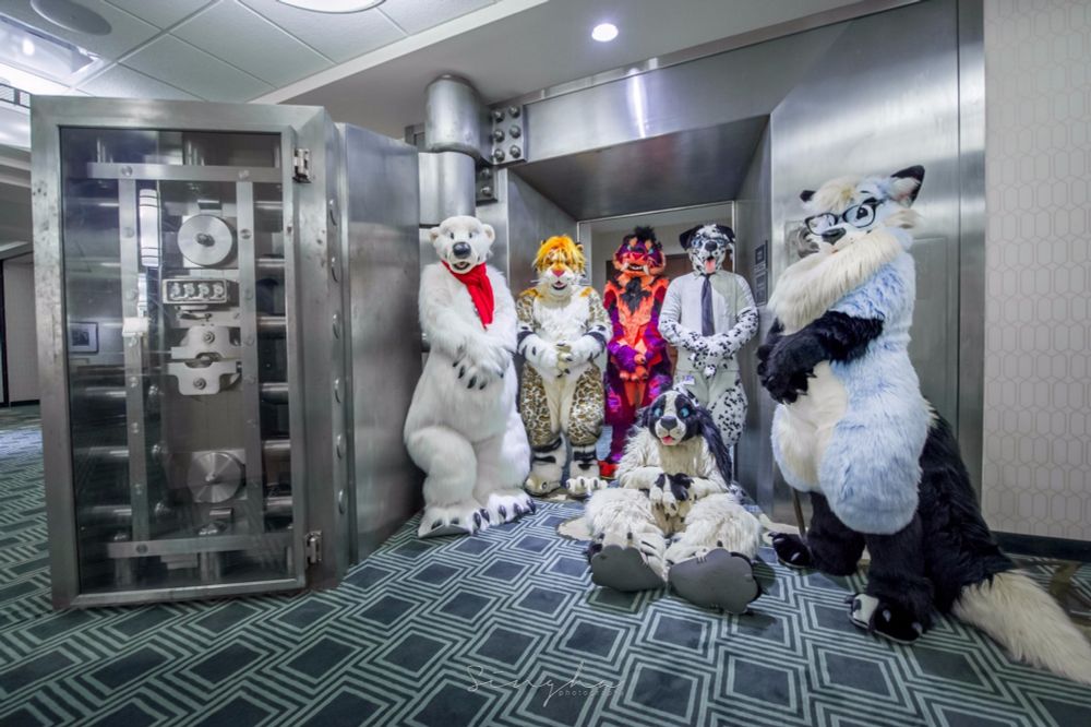 6 fursuits posting in front of a bank vault door a polar bear, 2 Sabrecats, a Dalmatian, an arctic fox and a dog (maybe cocker spaniel?) sitting on the floor