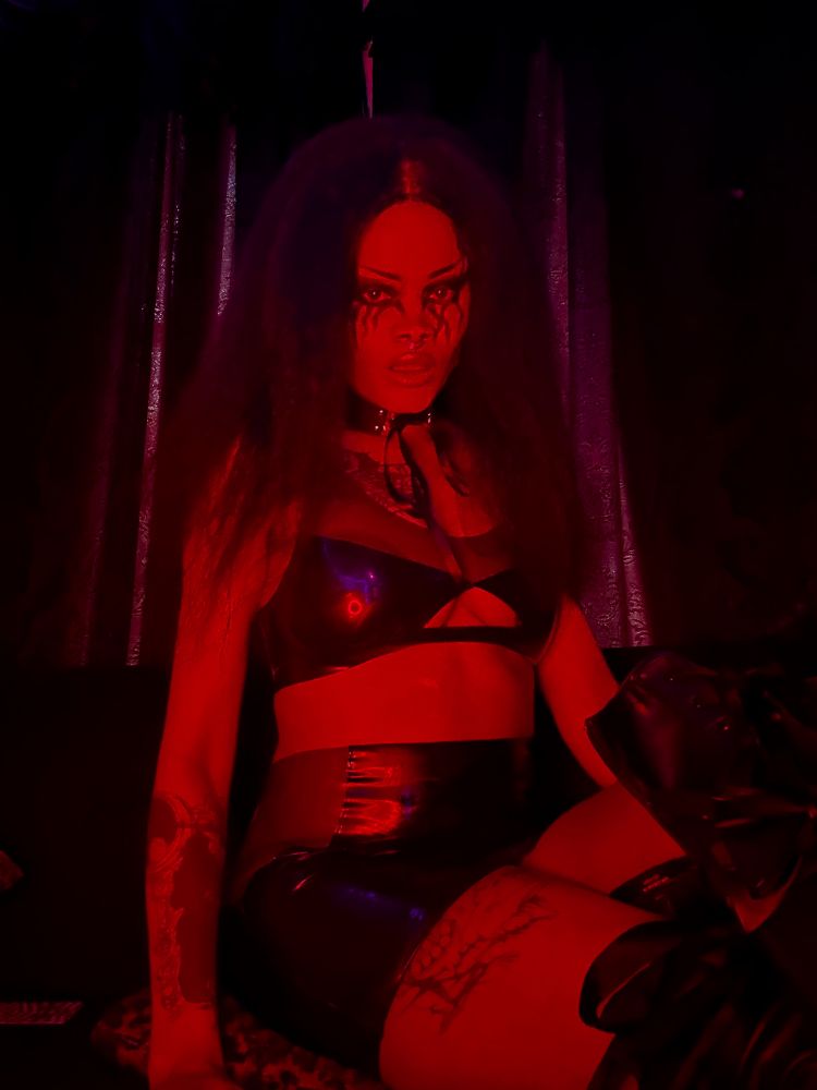 Domina Laveau is sitting on a couch with leopard print pillows in front of curtains. They are wearing metal makeup and shiny latex and a choker.
Save