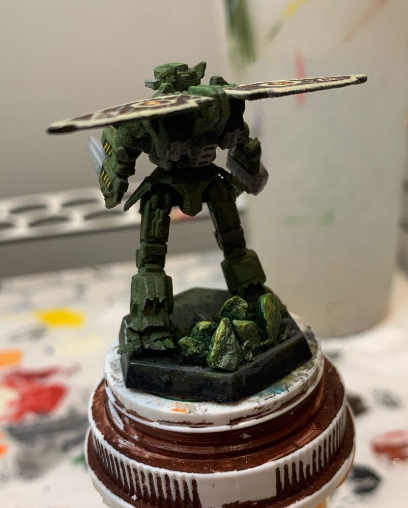 a picture of the agrotera from behind, this time emphasizing the dark green rocks on the base of the mini. the rocks are highlighted and dry brushed with light green and gold