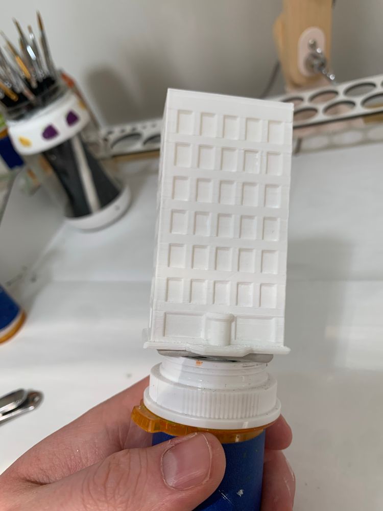 a 3d printed apartment building primed white. it stands suck to a pill bottle serving  as a paint handle