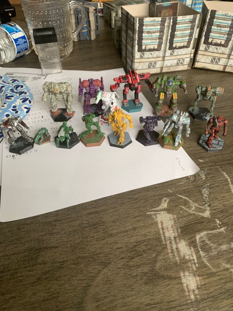 an assortment of 13 battletech minis painted in various schemes. an emperor painted like a marble statue covered in vines and flowers. an awesome painted in emulation of synth wave colors, purple drybrushed with orange. an archangel painted red with circuit lines on it to emulate the vector heart style. a mackie painted green and drybrushed with a rust color. an argus painted a desaturated green with orange stripes. an icarus II painted silver with a raven painted on its right shoulder and free painted on its left arm. a quad procyon protomech pained deep green, crouching to pounce. a boggart protomech painted the same green with hazard stripes on its sides. a flea painted light green with hazard stripes. a starslayer painted construction vehicle yellow. a fire falcon painted deep purple. a chameleon painted silver with stripes of light green and blue to make it look like it's shimmering with stealth armor. a pack hunter painted a deep blood red to look like v2 from ultra kill. and last but not least, an urbanmech LAM painted in white, but it's panel lines are painted a bright red on its body and green on its gun. 