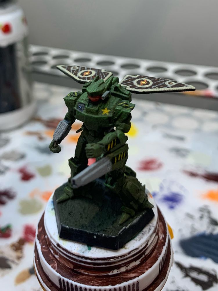 an agrotera form battletech painted a deep forest green, with bright green serving as a highlight. it is facing towards the left side of the screen. the agrotera comes equipped with a winged helmet shaped cockpit, a partial wing system on the back, a gauss rifle on the left arm (right from the viewers perspective, and some medium lasers on the right arm (left from the viewers perspective). each of the weapons is painted with a bright metalic gray. visible from this angle is the arotera's wings, which will be described in detail
later, which are painted like moth wings. on the left breast (right from the viewers perspective) is a simple gold star. on the right breast (left from the viewers perspective) is a small grey square that has a light blue talon painted in it. the visor of the cockpit is also painted a bright metallic orange, with bright metallic pink being used as a highlight color. also on the left arm two hazard stripes are painted on the forearm and the gauss rifle. these stripes are also painted on the shoulder pads of the mini. these stripes appear old and faded 