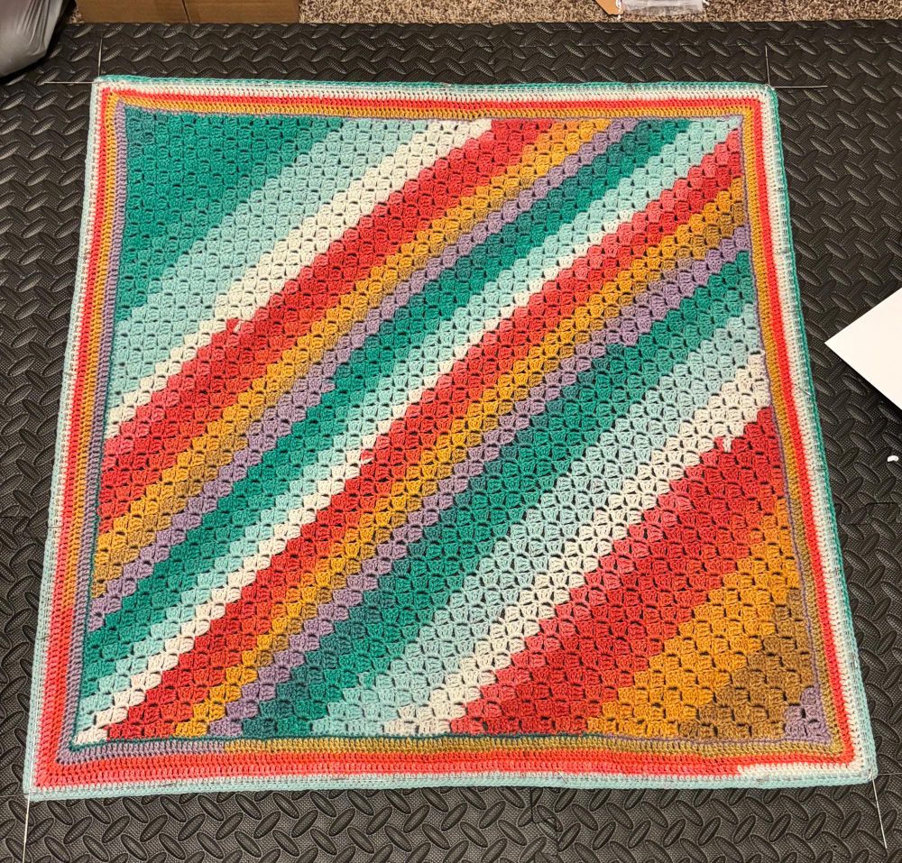 A recently completed, square, crocheted baby blanket, in diagonal stripes of colors with a bright, beachy feel is pinned out with blocking wires to dry.