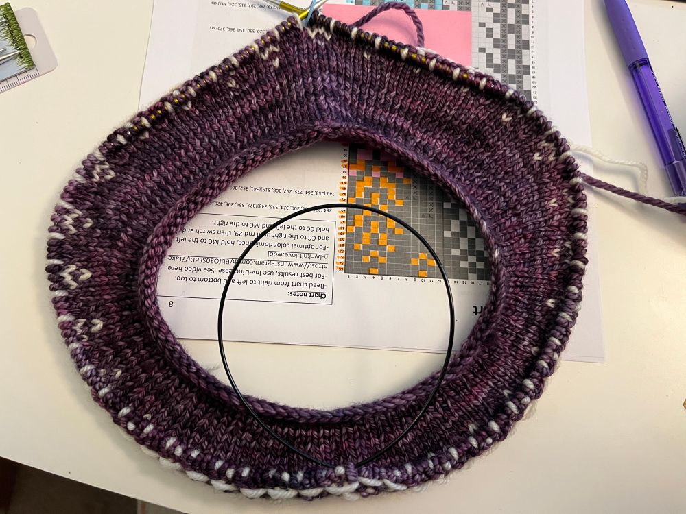 The neck of a knit sweater on circular needles. It’s made of purple yarn with the start of a white pattern.