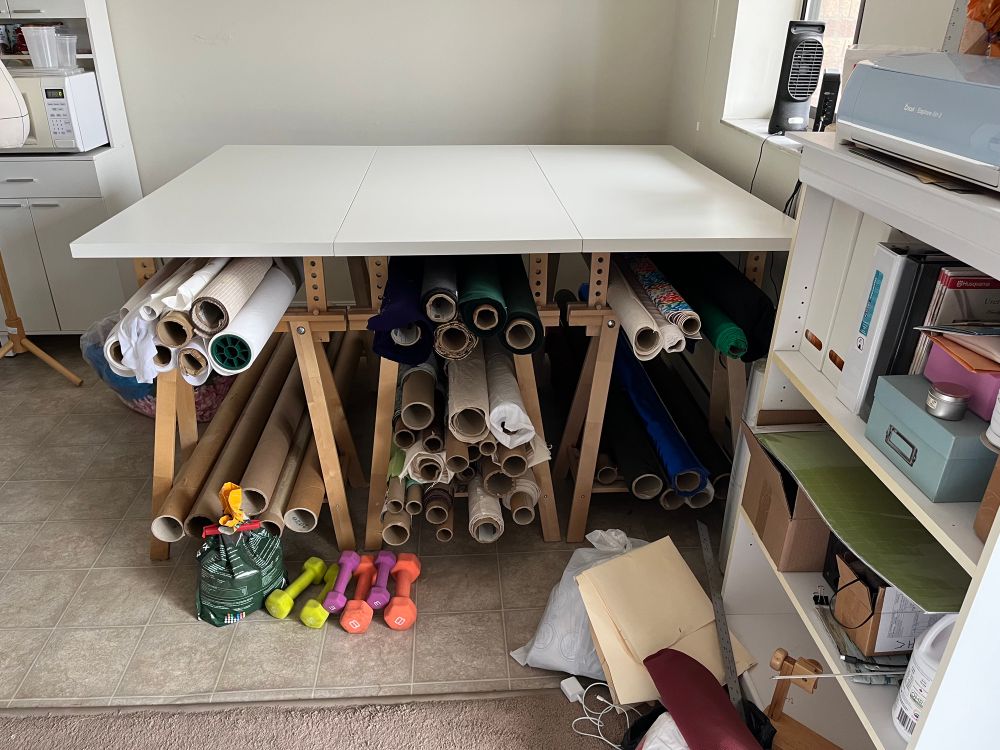 Three ikea trestle leg desks bolted together with rolls of fabric stored in the trestle legs sorted by type.
