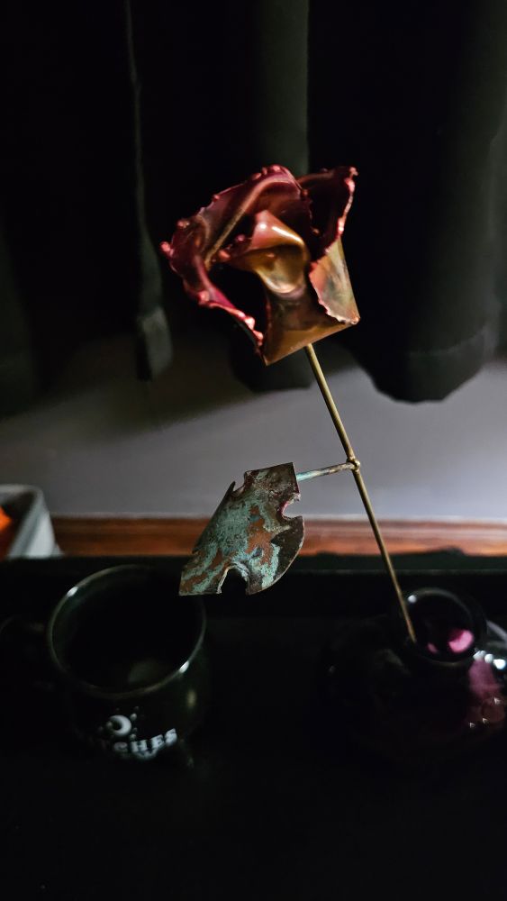 A copper rose with different colors including more copper tones on the base and pink tones at the petal tips where its been heated to different temperatures. The rose stem has one leaf. It is green from patina 