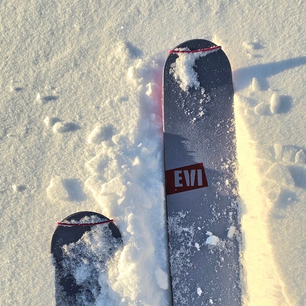 Skitupper (sølv) med "EVI" på rødt rektangel i outline i tørr løssnø og lav sol fra V. Tekst reklame for vår lokale skimaker Endre Hals som produserer EVI ski. Navnet henspeiler på at de kan repareres og resirkuleres. 