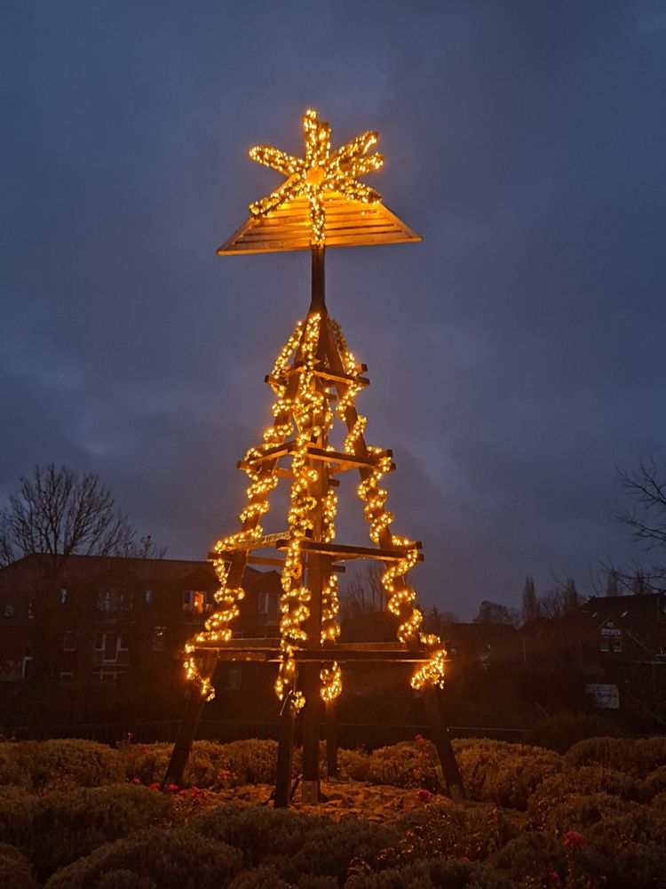 Ein hohes, kegelförmiges Holzgestell, das mit Lichterketten umwickelt ist mit einem sich auf der Spitze befindlichen beleuchteten Stern. Es ist noch dunkel und der Himmel ist bewölkt.