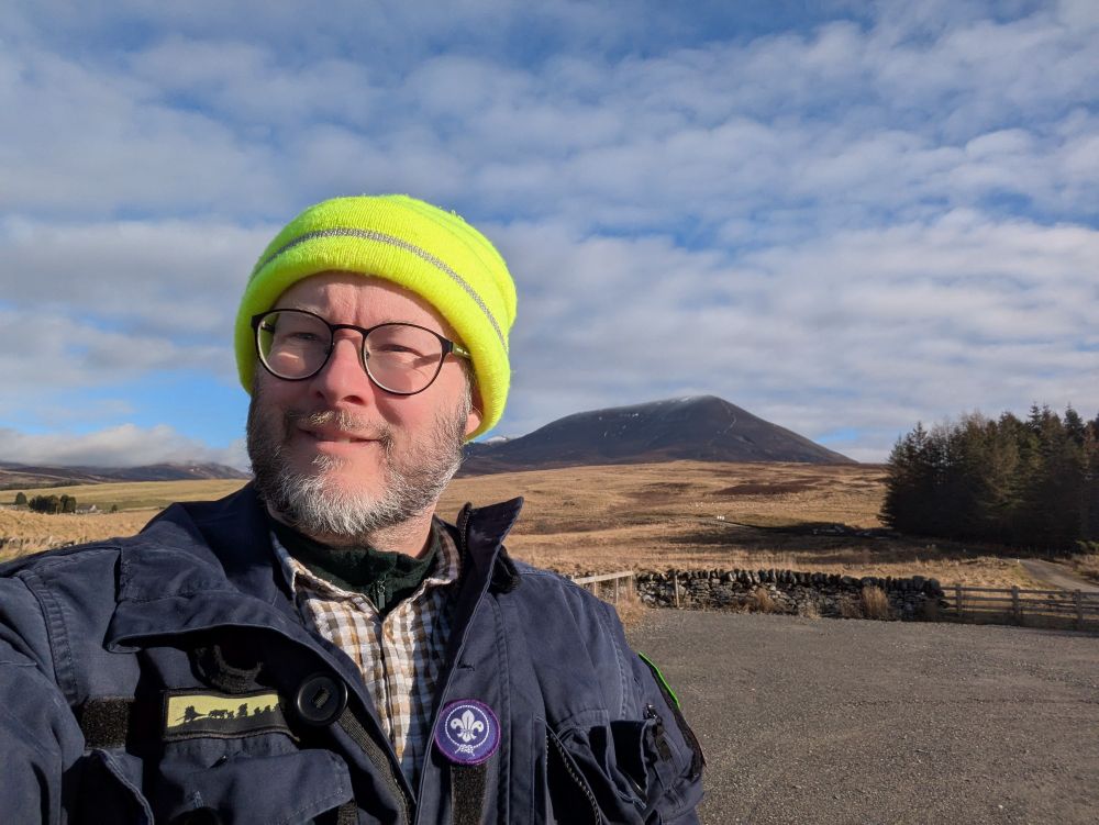 Me in my mountain walking jacket and fluorescent yellow high Vis hat. In the background is the mountain Càrn Liath, which is pretty bare with an obvious track all the way to the top. 