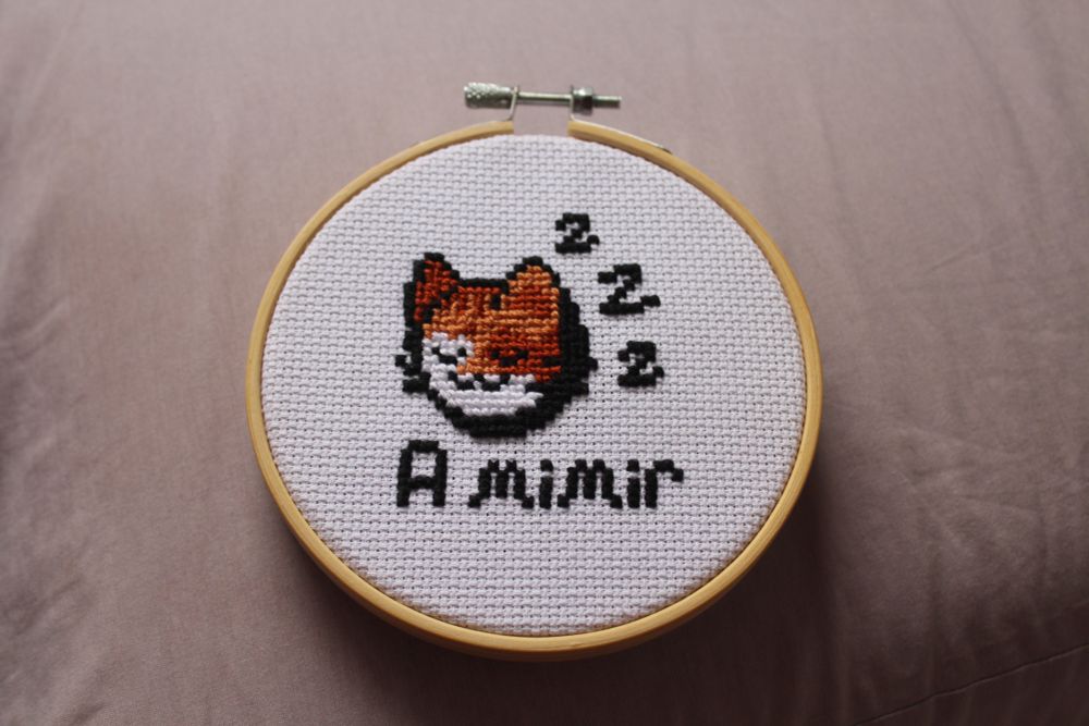 bordado em ponto-cruz num bastidor redondo de 12 cm. Bordei a cabeça de um gatinho de pelagem laranja, marrom e branca. O gatinho está de olhos fechados, tranquilo, ao seu lado há 3 letras "Z" indicando que está dormindo, e embaixo escrito: A mimir.