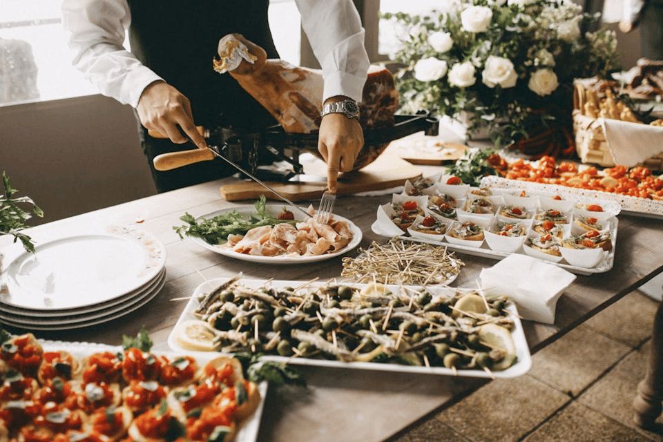 A well-dressed server is slicing cured meat at a lavish buffet table filled with an array of artfully presented appetizers. The spread includes trays of skewered olives with lemon slices, tomato-topped bruschetta, and small paper bowls filled with seafood and vegetable bites. A stack of clean white plates sits ready for guests, while a large floral arrangement and baskets of bread add elegance to the rustic wooden table. Natural light filters in, suggesting a festive and upscale gathering, perhaps a wedding or formal reception.