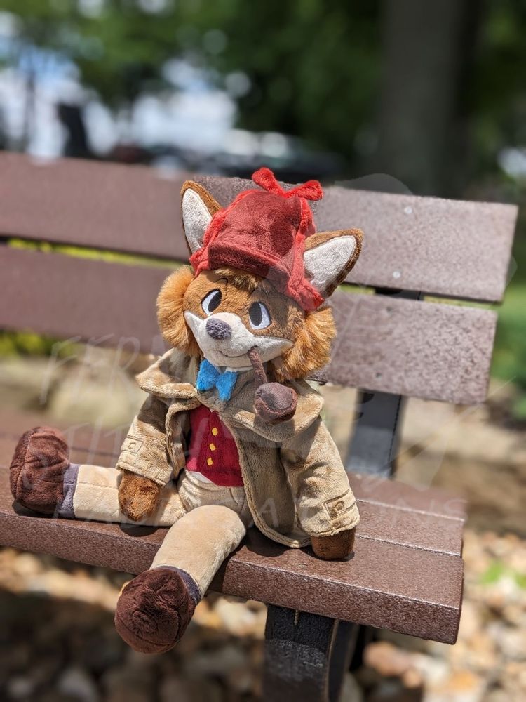 A photo of a handmade Sherlock Hound plush sitting on a park bench on a nice day. He's dressed in his standard fare: a red and burgundy deerstalker cap; a red vest; a blue bowtie; dark brown shoes; and a tan overcoat. His pipe hangs from his lips, which are quirked up in his trademark half-smile. One of his legs is stretched out across the bench, while the other dangles over the edge. He's leaning back on one arm.