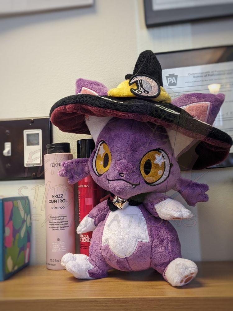 A photo of mauve chibi cat plush with golden eyes, a white belly, and white socks. It's wearing a black witch hat with a burgundy underside and a yellow ribbon adorned with a buckle featuring a pair of shears inside a crescent moon. Around its neck is a black cape and a silver bell affixed with a burgundy cord.