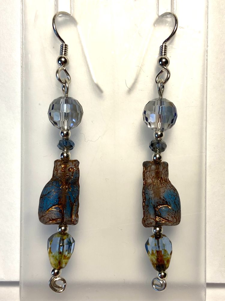 These sterling silver earrings have three large glass beads and one tiny one, separated by little round sterling silver beads. The bead at the top is a faceted crystal round in clear glass with grey-blue-purple finish. Below it is a tiny faceted Swarovski crystal bead in black diamond colorway, with aurora borealis. Next down is the biggest bead, a Czech glass cat in blue and clear, with bronze accents. At the bottom is an inverted faceted teardrop of blue and amber glass.