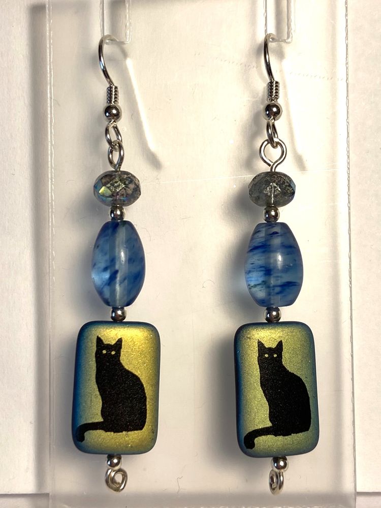 These sterling silver earrings have a stack of three beads. The smallest, at the top, is a faceted Czech glass rondelle with aurora borealis finish which makes it look aqua with flashes of blue and purple. The middle bead is a slightly larger pointed cylinder (a big rice bead) of clear glass with wisps of blue glass trailed through it. The bottom bead is a larger tablet bead, rectangular with rounded corners, of black glass with aurora borealis finish, etched to show a black cat on both sides.