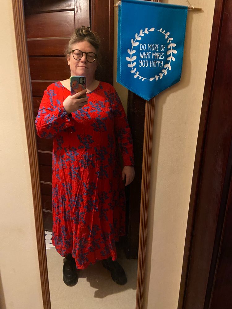 A fat white person in a long red cotton dress with blue flowers  is taking a selfie next to a turquoise banner with a white laurel wreath that says DO MORE OF WHAT MAKES YOU HAPPY. The person looks sleepy. Their hair is piled into an untidy updo. They are wearing black Fluvog boots, one of which has a thick prosthetic sole.