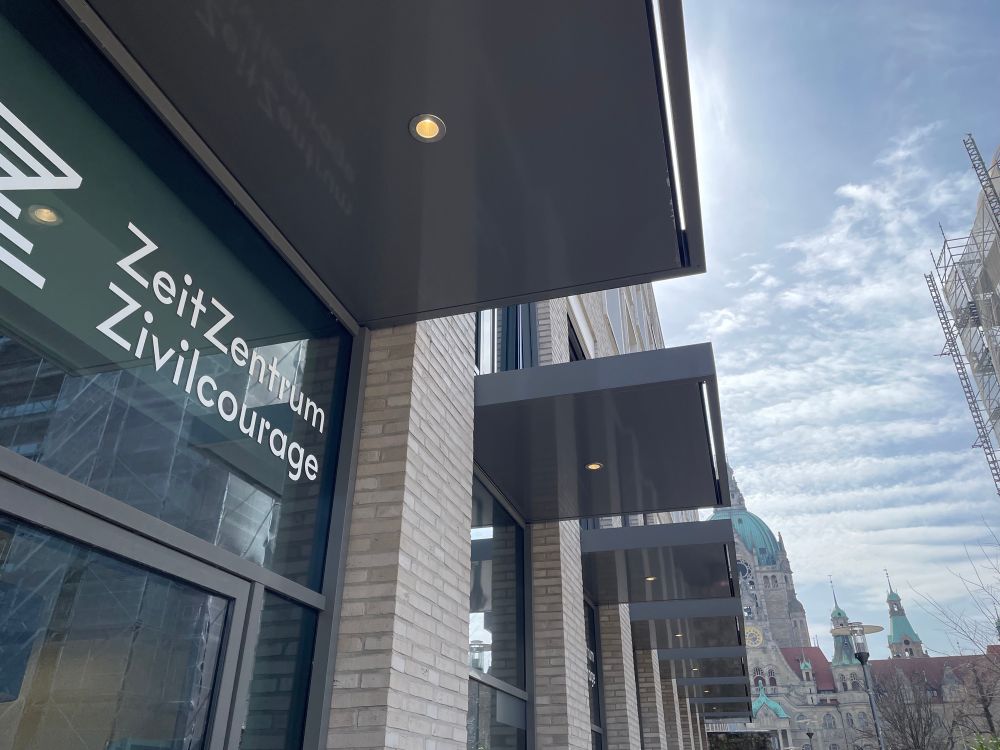 Ein Foto von einem Gebäude im Vordergrund. Man erkennt den Schriftzug „ZeitZentrum Zivilcourage“. Im Hintergrund zu sehen ist das Rathaus von hannover. 