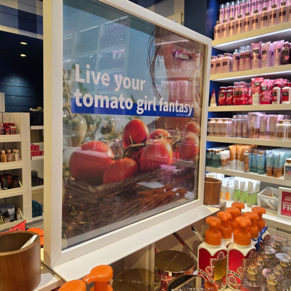 Bath and bodyworks display that says "Live your tomato girl fantasy" with a white woman smiling over a basket of tomatoes. Its to advertise a tomato scented spray and hand soap.