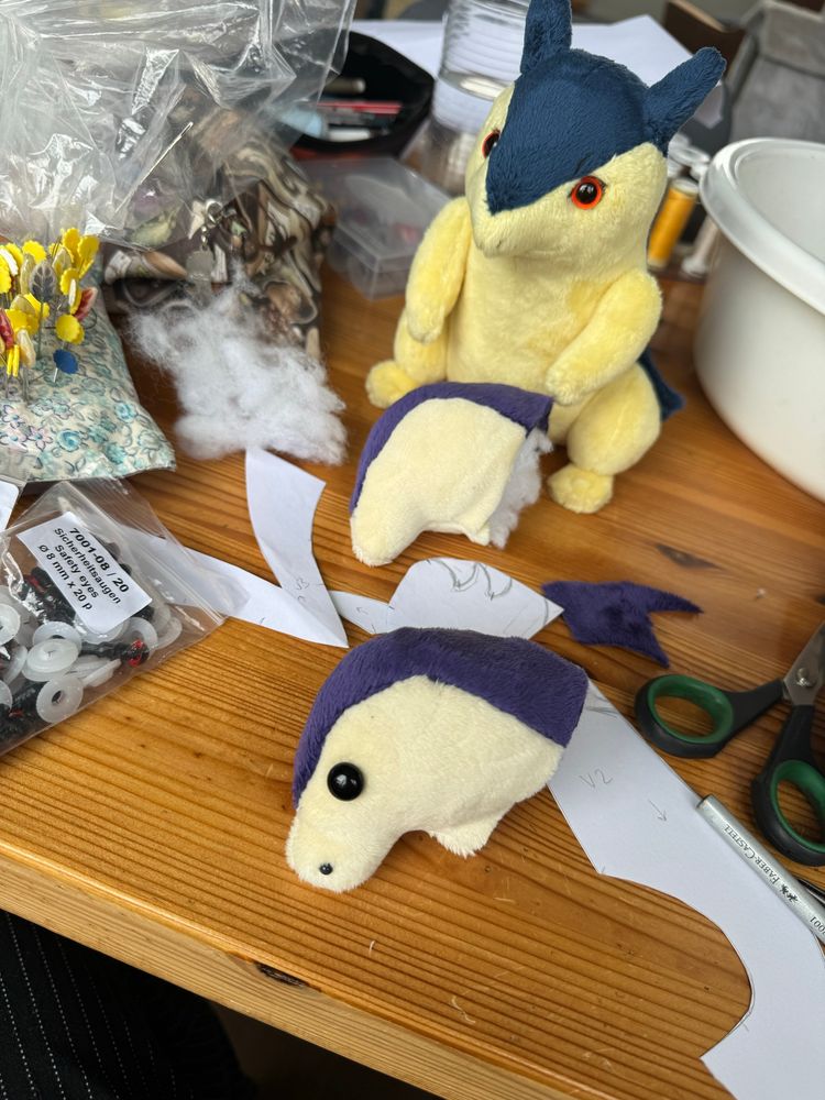 Photo of a work desk with various sewing supplies, two loose plush heads and one finished typhlosion plush looking slightly concerned 