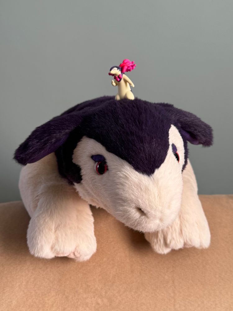 A photo of a large realistic plush Hisuian typhlosion’s head and front paws facing the camera. A tiny plastic figurine of a Hisuian typhlosion is standing on its head. 