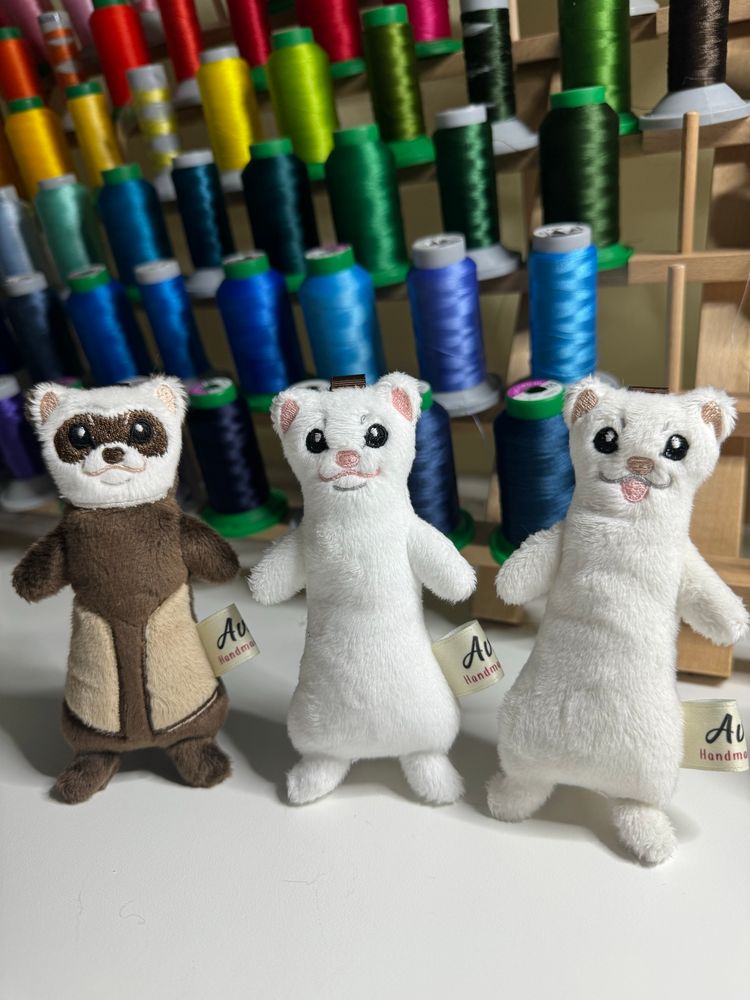 Photo of 3 small ferret plushies, two white and one brown, standing in front of a rack of rainbow threads. 