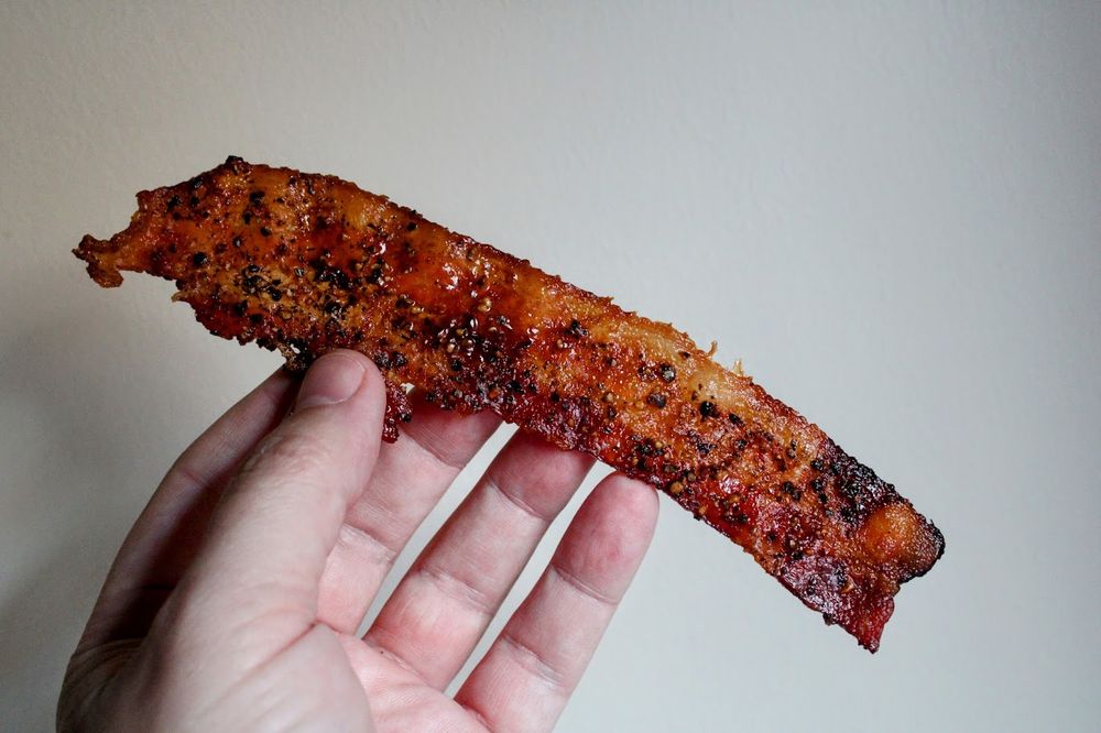 A hand holds a piece of bacon that has lots of ground black pepper on it.
