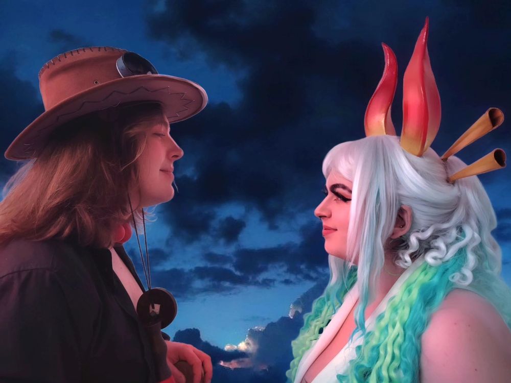 An edited photo of Dan cosplaying Yamato and his friend cosplaying Ace from One Piece. They are facing each other and smiling. They are illuminated by a warm light. The background is a dark blue cloudy sky.
