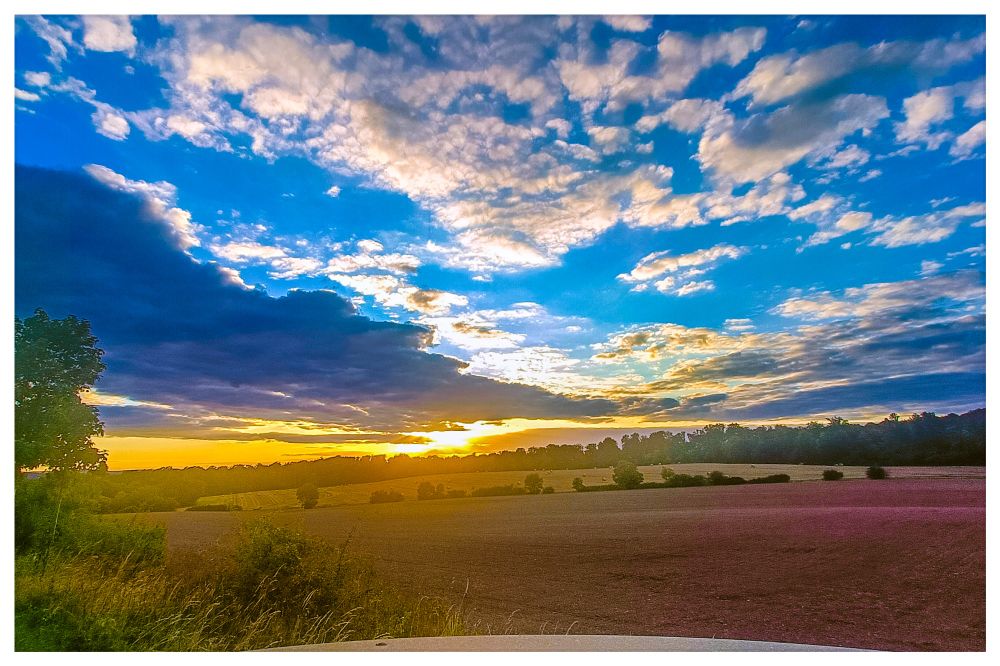 Landschaftsaufnahme, Feld mit Strohballen, Wald am Horizont, Himmel mit Wolken, Sonnenstrahlen brechen mittig durch die Wolken. Per Farbfilter wurde die Farben verstärkt. 