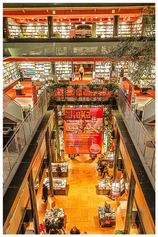 Das Bild zeigt das Innere einer großen, mehrstöckigen Buchhandlung. Mehrere Etagen mit Regalen voller Bücher sind offen einsehbar. In der Mitte hängt ein großes rotes Werbebanner. Menschen bewegen sich zwischen den Tischen und Regalen, einige stöbern in Büchern. Die Atmosphäre ist hell, warm beleuchtet und belebt.