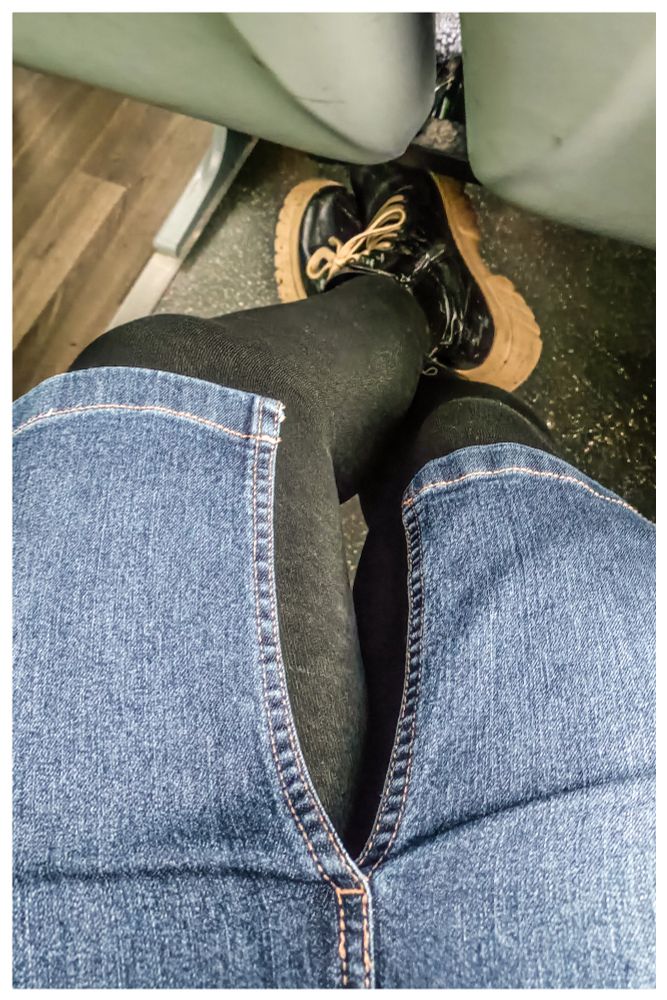 Frauenbeine, leicht überschlagen, schwarze Strumpfhose, Jeansrock mit Schlitz vorne, schwarze Stiefel mit heller Schnürung und Sohle. In einem Bus aufgenommen, ein Stück des Vordersitzes ist sichtbar. 