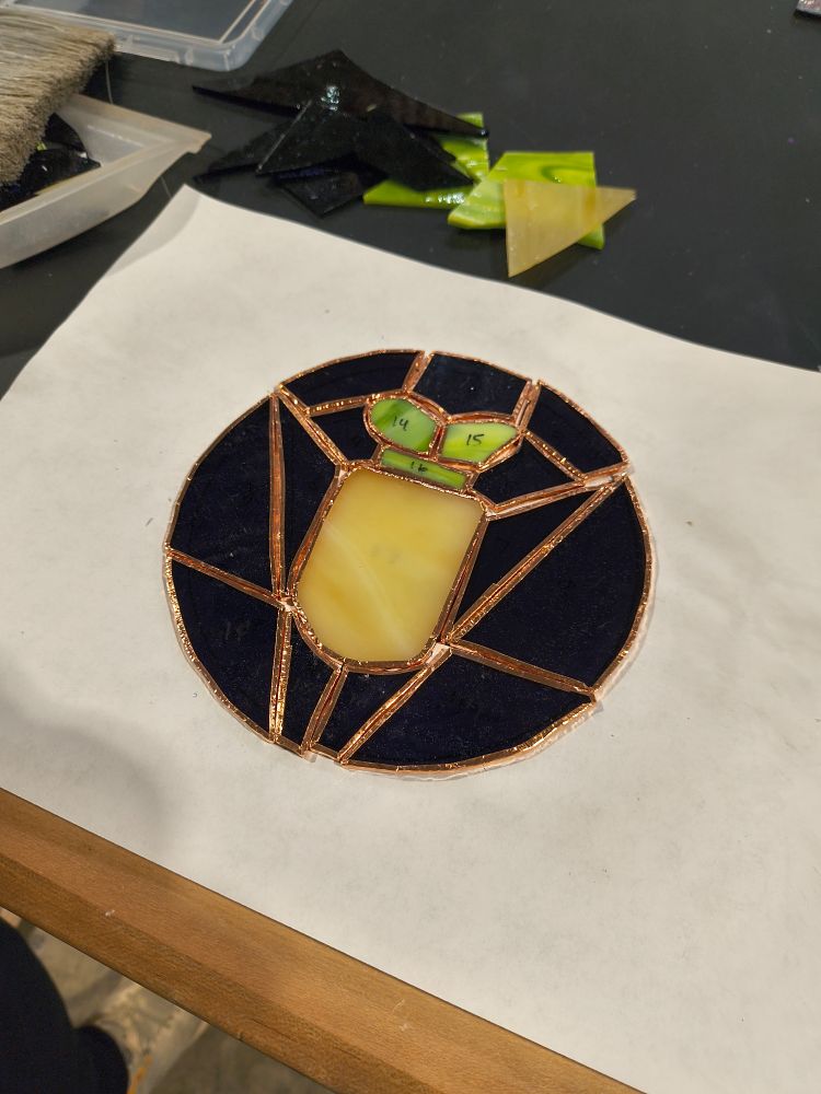 A photo of the same glass pieces, now with copper foil and arranged into a rough shape of a radish with green leaves on a dark purple background 