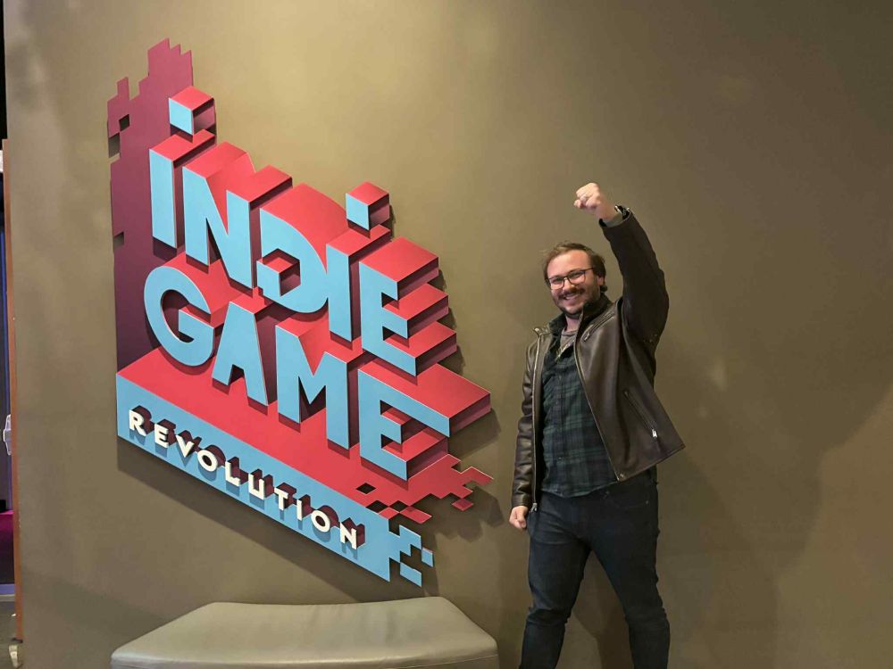 A photo of a man with glasses and a beard, raising his fist next to a sign which reads "Indie Game Revolution" in stylized red and blue text. The man is wearing a leather jacket over a green flannel shirt